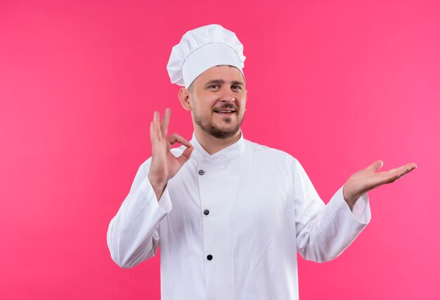 Smiling young handsome cook in chef uniform doing ok sign and showing empty hand isolated on pink space