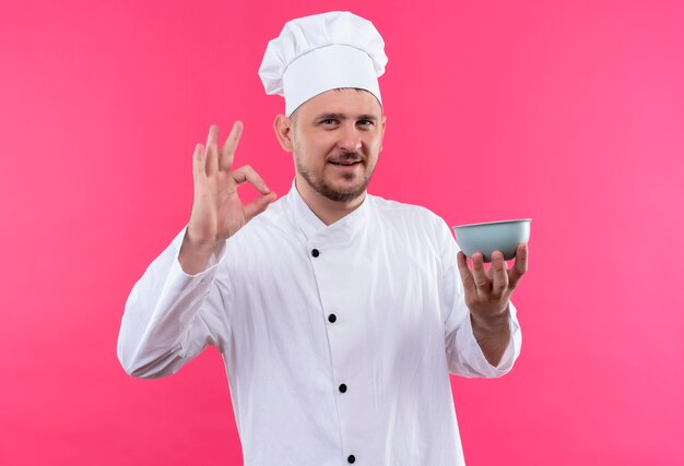 Улыбающийся молодой красивый повар в униформе шеф-повара, держащий миску и делающий знак ОК, изолирован на розовом пространстве