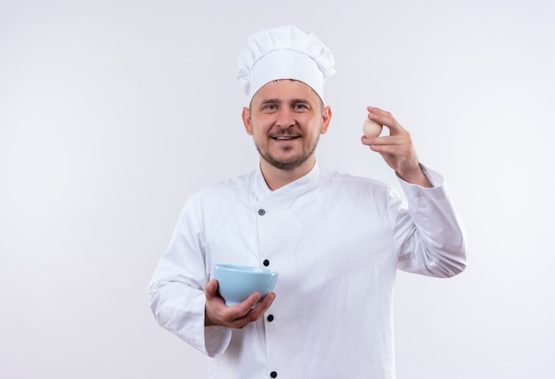 Smiling young handsome cook in chef uniform holding bowl and egg isolated on white space