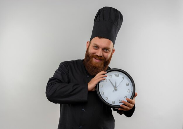 白いスペースに孤立して見える時計を保持しているシェフの制服を着た若いハンサムな料理人の笑顔