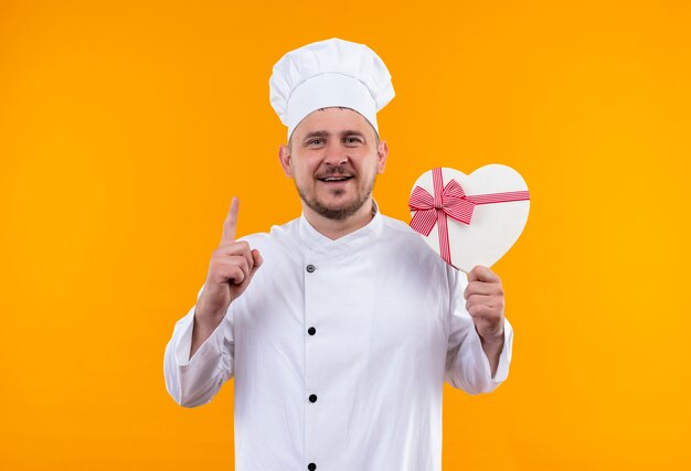 Smiling young handsome cook in chef uniform holding heart shaped gift box and raising finger on isolated orange space