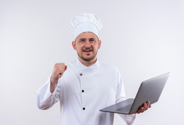 Smiling young handsome cook in chef uniform holding laptop and raising fist on isolated white space