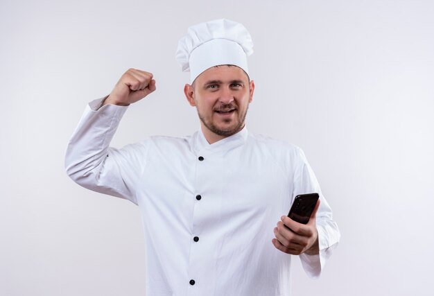 Smiling young handsome cook in chef uniform holding mobile phone and doing strong gesture isolated on white space
