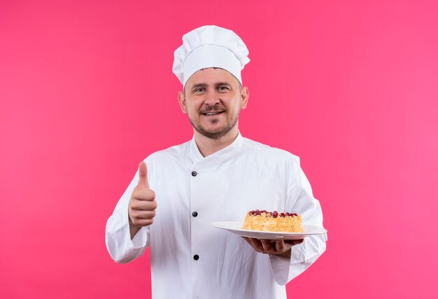 Smiling young handsome cook in chef uniform holding plate of cake showing thumb up isolated on pink space