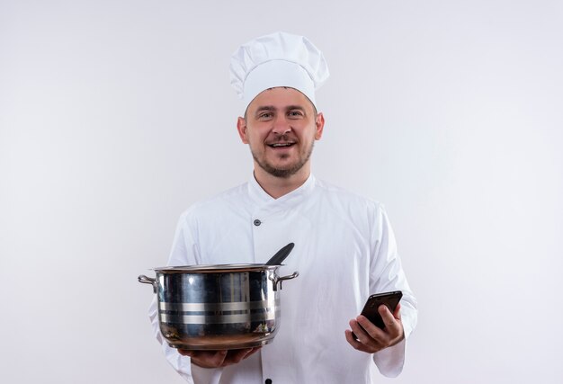 Smiling young handsome cook in chef uniform holding pot and mobile phone on isolated white space 