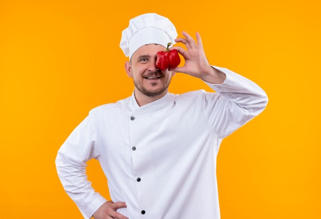 Улыбающийся молодой красивый повар в униформе шеф-повара кладет перец на глаза, изолированные на оранжевом пространстве