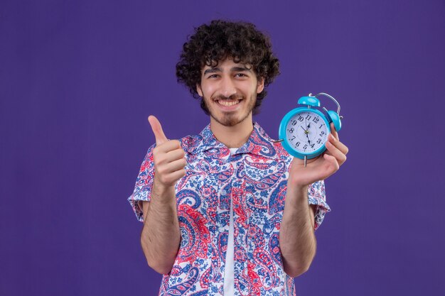 Smiling young handsome curly traveler man holding alarm clock showing thumb up on isolated purple wall with copy space