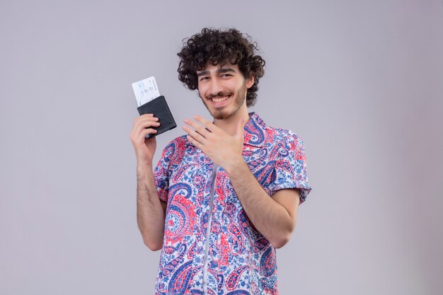 Smiling young handsome curly traveler man holding wallet and airplane tickets and pointing with hand at it on isolated white wall with copy space