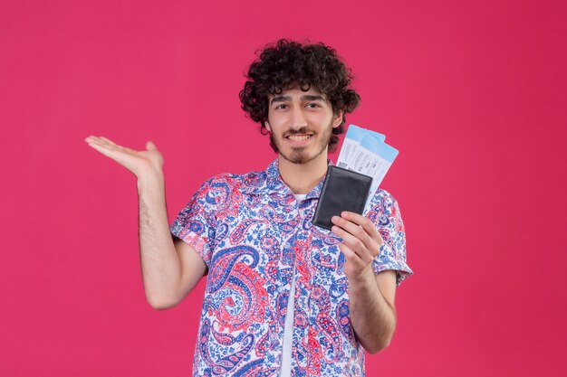 Smiling young handsome curly traveler man holding wallet and airplane tickets and showing empty hand on isolated pink wall with copy space