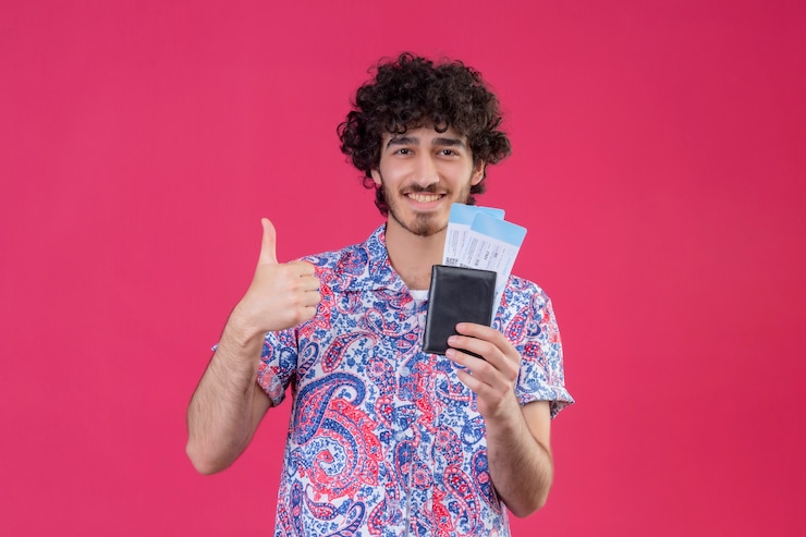 Free Photo smiling young handsome curly traveler man holding wallet and airplane tickets and showing thumb up on isolated pink wall with copy space