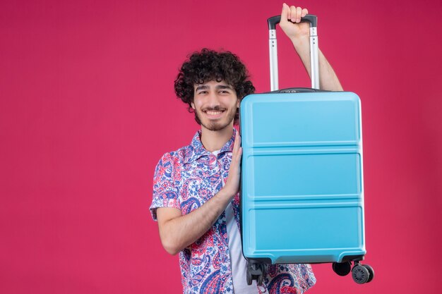 Smiling young handsome curly traveler man raising suitcase and putting hand on it on isolated pink wall with copy space