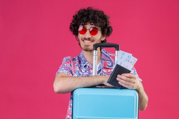 Smiling young handsome curly traveler man wearing sunglasses holding wallet and airplane tickets looking at left side with hands on suitcase on isolated pink wall with copy space