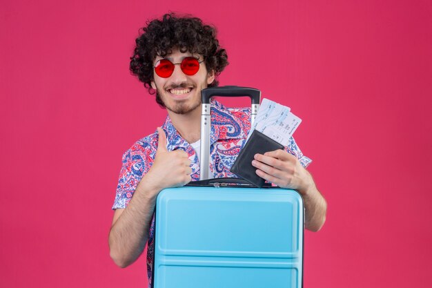 Smiling young handsome curly traveler man wearing sunglasses holding wallet and airplane tickets showing thumb up with suitcase on isolated pink wall with copy space