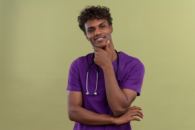 Улыбающийся молодой красивый темнокожий врач с кудрявыми волосами в фиолетовой форме со стетоскопом, держащий руку за подбородок в зеленой зоне