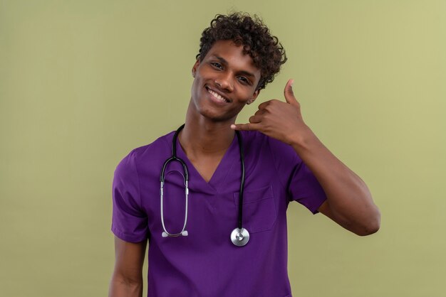 A smiling young handsome dark-skinned doctor with curly hair wearing violet uniform with stethoscope holding hand near ear gesture telephone sign with fingers on a green space