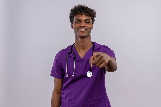 A smiling young handsome dark-skinned doctor with curly hair wearing violet uniform with stethoscope pointing at camera with index finger 