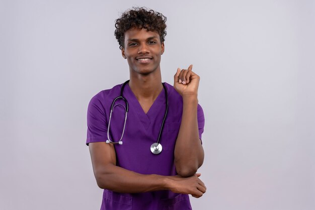 A smiling young handsome dark-skinned doctor with curly hair wearing violet uniform with stethoscope pointing with index finger while  