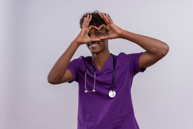 A smiling young handsome dark-skinned doctor with curly hair wearing violet uniform with stethoscope showing heart shape sign with fingers 