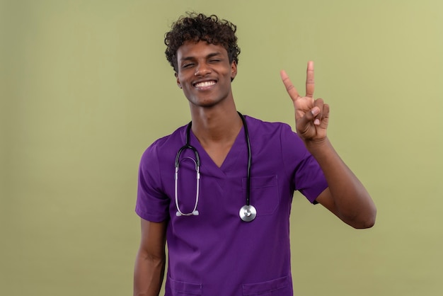 緑のスペースで2本の指を示す聴診器で紫の制服を着た巻き毛の笑顔の若いハンサムな浅黒い医者