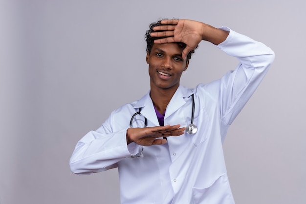 Улыбающийся молодой красивый темнокожий доктор с вьющимися волосами в белом халате со стетоскопом радостно смотрит в камеру