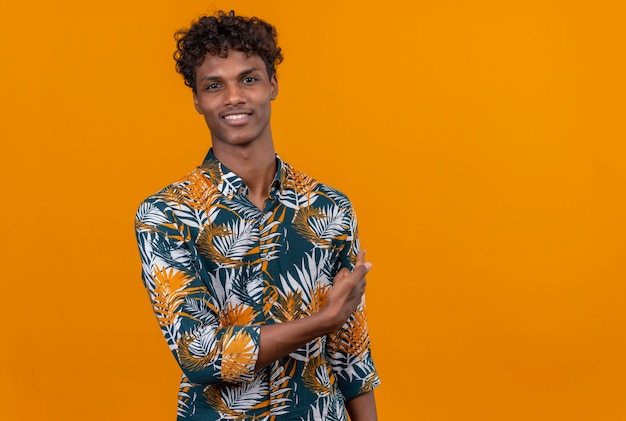 Smiling young handsome dark-skinned man with curly hair in leaves printed shirt showing something with hand on an orange background