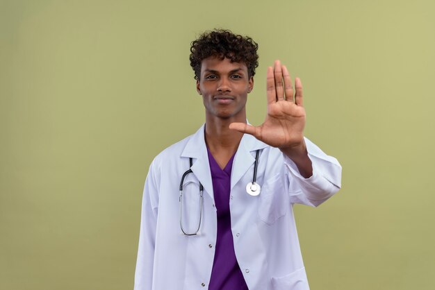 A smiling young handsome dark-skinned man with curly hair wearing white coat with stethoscope giving high five gesture on a green space