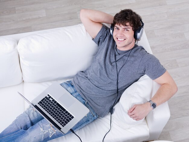 Smiling young handsome guy listening music in headphone from laptop - high angle