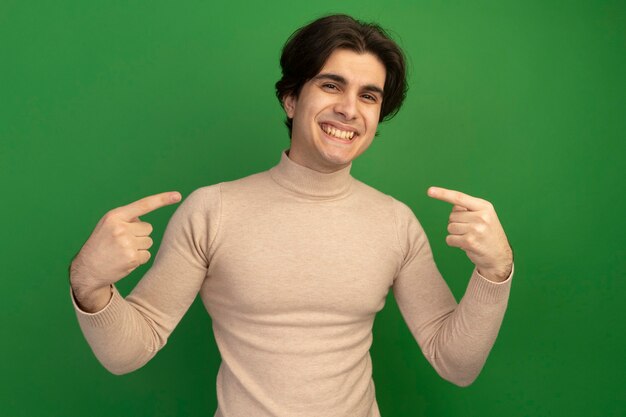 Smiling young handsome guy points at himself isolated on green wall