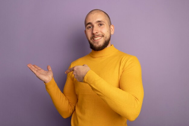 Smiling young handsome guy pretending holding and points at something isolated on purple