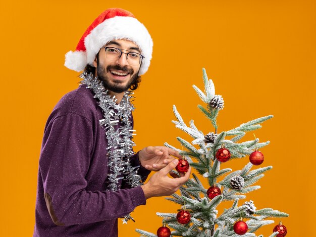 Smiling young handsome guy standing nearby christmas tree wearing christmas hat with garland on neck isolated on orange background