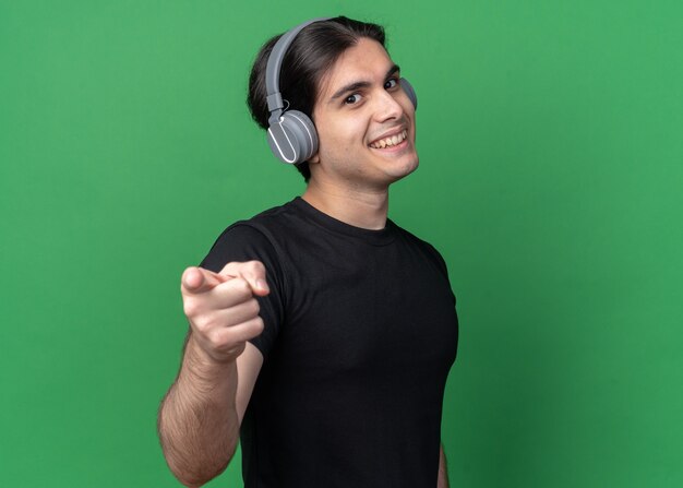 Smiling young handsome guy wearing black t-shirt and headphones showing you gesture isolated on green wall