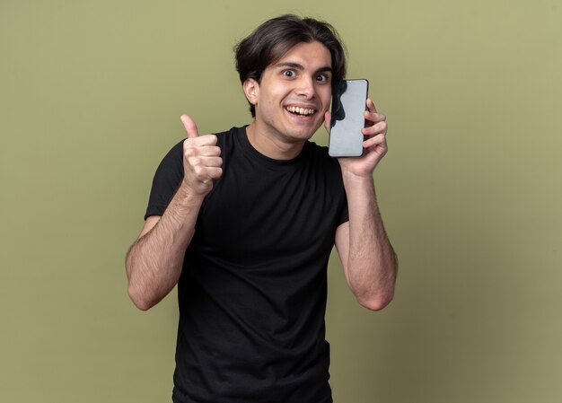Smiling young handsome guy wearing black t-shirt holding alarm clock around face showing thumb up isolated on olive green wall