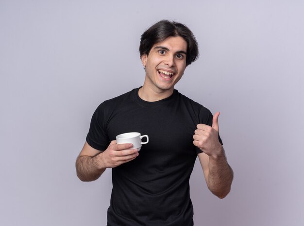 Smiling young handsome guy wearing black t-shirt holding cup of coffee showing thumb up isolated on white wall