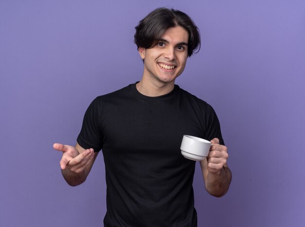 紫色の壁に分離されたジェスチャーを示すコーヒーのカップを保持している黒いTシャツを着て笑顔の若いハンサムな男