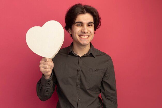 Smiling young handsome guy wearing black t-shirt holding heart shape box isolated on pink wall