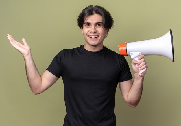 Smiling young handsome guy wearing black t-shirt holding loudspeaker and spreading hand isolated on olive green wall