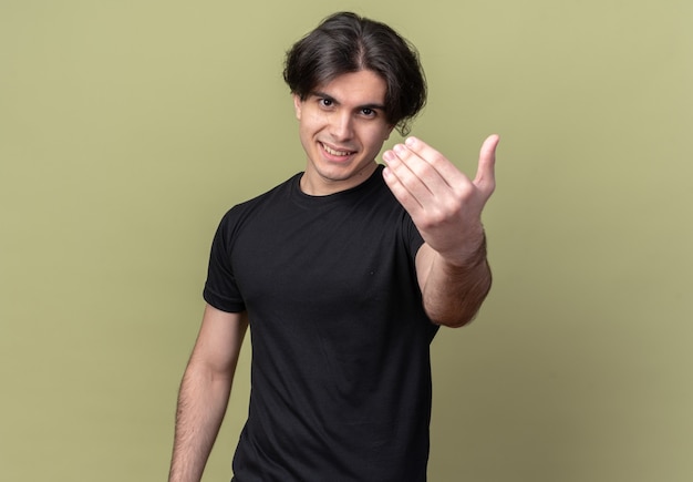 Smiling young handsome guy wearing black t-shirt holding out hand at front isolated on olive green wall