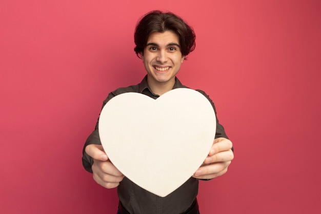 Улыбающийся молодой красивый парень в черной футболке, протягивая коробку в форме сердца спереди, изолированную на розовой стене