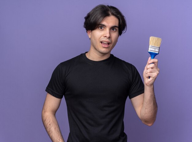 Smiling young handsome guy wearing black t-shirt holding paint brush isolated on purple wall