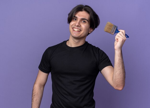 Smiling young handsome guy wearing black t-shirt holding paint brush isolated on purple wall