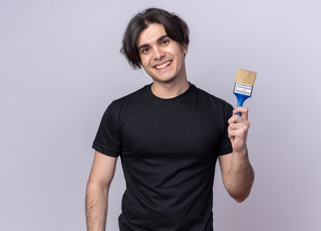 Smiling young handsome guy wearing black t-shirt holding paint brush isolated on white wall