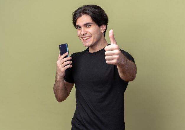 コピースペースとオリーブグリーンの壁に分離された親指を示す電話を保持している黒いTシャツを着て笑顔の若いハンサムな男