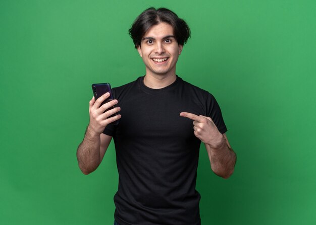 Smiling young handsome guy wearing black t-shirt holding and points at phone isolated on green wall
