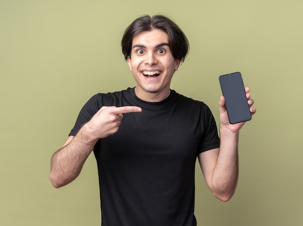 Улыбающийся молодой красивый парень в черной футболке держит и указывает на телефон, изолированный на оливково-зеленой стене