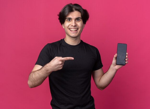 Smiling young handsome guy wearing black t-shirt holding and points at phone isolated on pink wall