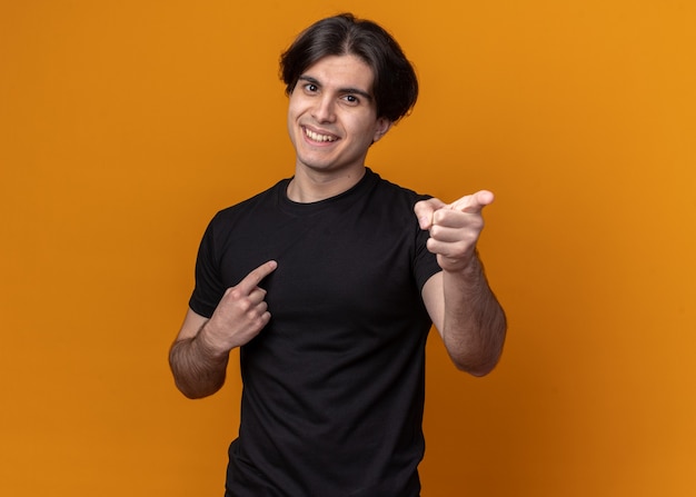 Smiling young handsome guy wearing black t-shirt points at side isolated on orange wall with copy space