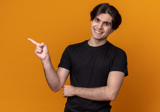 Smiling young handsome guy wearing black t-shirt points at side isolated on orange wall with copy space