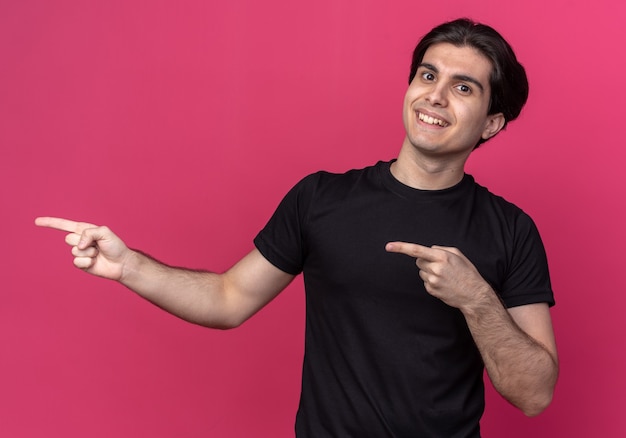 Smiling young handsome guy wearing black t-shirt points at side isolated on pink wall with copy space