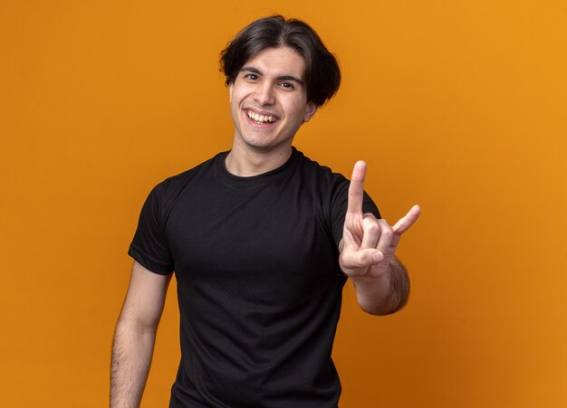 Smiling young handsome guy wearing black t-shirt showing goat gesture isolated on orange wall
