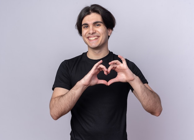 Smiling young handsome guy wearing black t-shirt showing heart gesture isolated on white wall